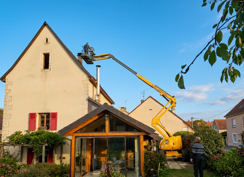 Un technicien chauffagiste BIO'CHAUF en tenue professionnelle (combinaison ou polo de marque) effectuant une intervention de maintenance ou dépannage sur une chaudière ou un système de chauffage à l'intérieur d'un domicile. Le technicien est à genoux ou penché, examinant ou réparant l'équipement avec ses outils. La scène se déroule dans un sous-sol ou une chaufferie résidentielle, illustrant un service après-vente professionnel et fiable directement assuré en interne.